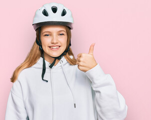 Beautiful young caucasian girl wearing bike helmet smiling happy and positive, thumb up doing excellent and approval sign