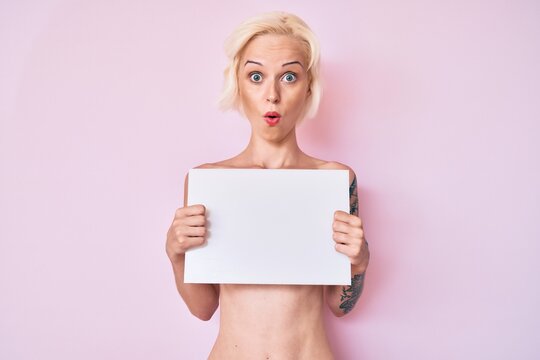 Young blonde woman with tattoo standing shirtless holding empty blank banner scared and amazed with open mouth for surprise, disbelief face