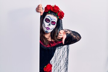 Young woman wearing day of the dead custome holding blank empty banner looking unhappy and angry showing rejection and negative with thumbs down gesture. bad expression.