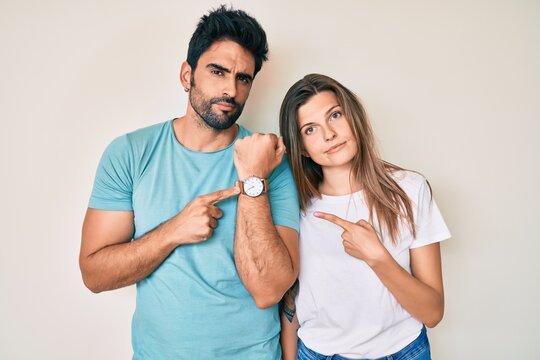 Beautiful Young Couple Of Boyfriend And Girlfriend Together In Hurry Pointing To Watch Time, Impatience, Looking At The Camera With Relaxed Expression
