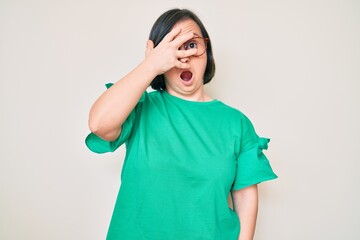 Brunette woman with down syndrome wearing casual clothes and glasses peeking in shock covering face and eyes with hand, looking through fingers afraid