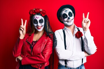 Couple wearing day of the dead costume over red smiling with happy face winking at the camera doing victory sign with fingers. number two.