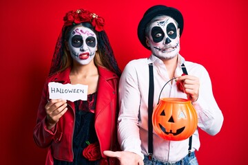 Couple wearing day of the dead costume holding pumpking and halloween paper clueless and confused expression. doubt concept.