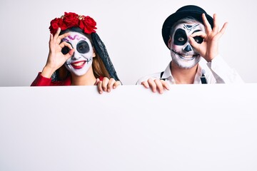 Couple wearing day of the dead costume holding blank empty banner smiling happy doing ok sign with hand on eye looking through fingers