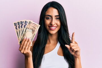 Beautiful hispanic woman holding japanese yen banknotes smiling with an idea or question pointing finger with happy face, number one