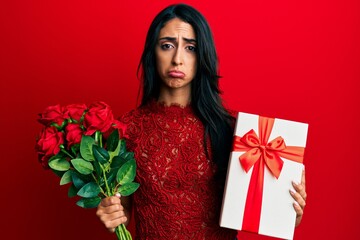 Beautiful hispanic woman holding anniversary gift and roses bouquet depressed and worry for distress, crying angry and afraid. sad expression.