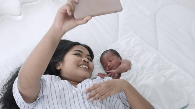 Close up shot of mother and her newborn baby making a selfie or video call to family on the bed white in home. Mixed race black boy Ethnicity Thai-Nigeria. New generation Technology Communication