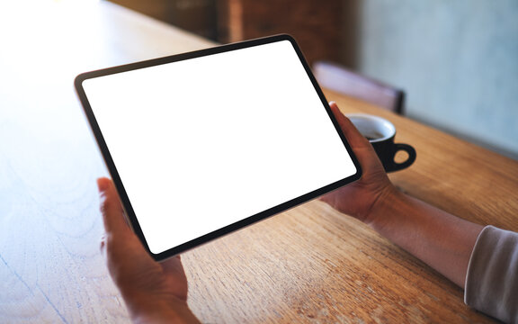 Mockup image of a woman holding digital tablet with blank white desktop screen with coffee cup on wooden table