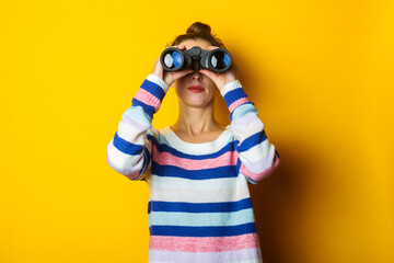 Young woman in sweater looking through binoculars on yellow background