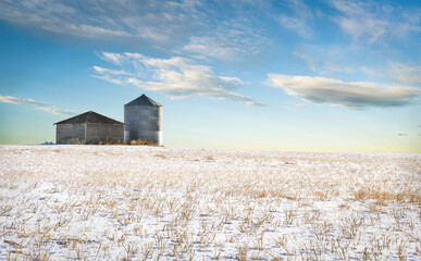 An empty grain silo and shed sit on a harvested snow covered field on the Canadian prairies in Rocky View County Alberta. - Powered by Adobe
