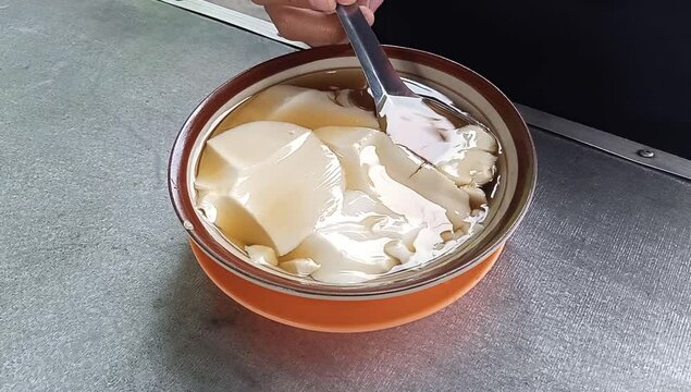 close up shot of chinese sweet traditional food called douhuwa tauwa bean curd tofu pudding with hot ginger syrup and showing hand scooping it