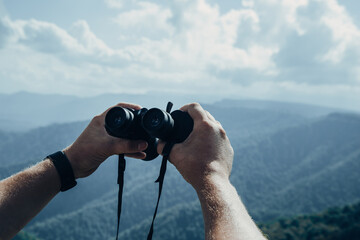 Hands holding binoculars on mountains forest nature background, looking through binoculars, travel, search and search concept.