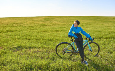 Fototapeta na wymiar A young cyclist in a tracksuit stopped in a field on a background of green grass in sunny weather.