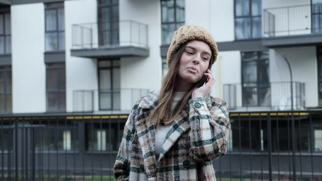 Cute and pretty girl with a beautiful appearance and stylish clothes talking on the phone with friends on the urban street background of new city building.