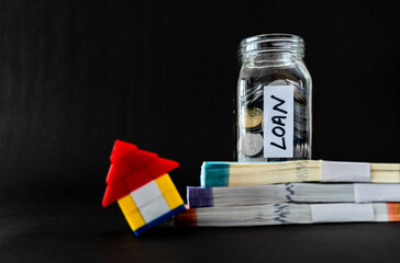 Glass jar on top of money bundles containing coins, with LOAN label on it and defocused leaning house toy.