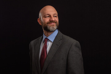 Portrait of a bald executive in a grey suit on a dark background. Male model in his 40s with grey beard. Looking away smiling.