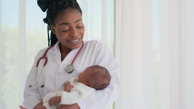 African American woman doctor hold sleeping newborn baby in her chest and look like she sing baby to sleep in front of white curtain in pediatric clinic or hospital with children healthcare concept.