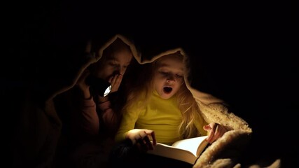 children reading a book under a blanket with light. two little girls is read a book under a blanket with a flashlight in a dark room at night, in bed.