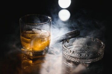 A glass of whiskey or Bourbon with ice cubes and a cigar on a black slate with smoke