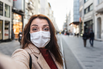 Beautiful girl wearing protective medical mask and fashionable clothes takes selfie with a smart phone at street. New normal lifestyle concept.