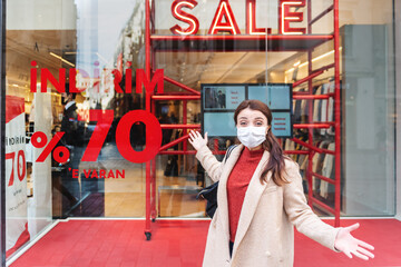 Beautiful girl wearing protective medical mask and fashionable clothes stands in front of sale,discount clothes store. New normal lifestyle concept.