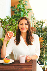Fototapeta na wymiar Young happy woman having breakfast on the balcony, eating grapefruit or red orange, cup of tee on table