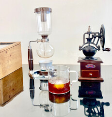 Japanese siphon vacuum coffee maker, iced espresso in a clear mug, grinder, and coffee bean storage wooden box on a clear, reflective glass table and a window curtain as a background.