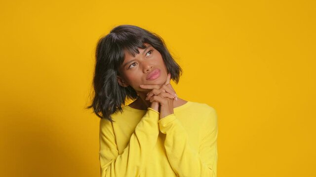 Pensive dreamy wavy haired dark skinned woman keeps hands under her chin looks around and thinks about something pleasant dressed in casual clothing isolated over gold yellow background. Let me think