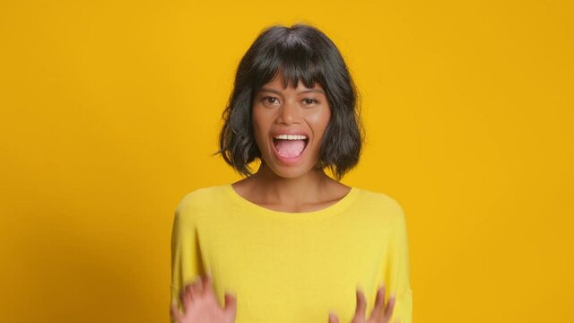 Emotive dark skinned young woman with excited face expression reacts on unexpected surprise raises hands hears great news shakes hands wears casual jumper isolated over gold yellow studio background