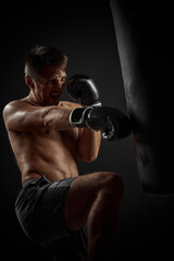 Fototapeta na wymiar muscular boxer in black boxing gloves punching in boxing bag on dark background