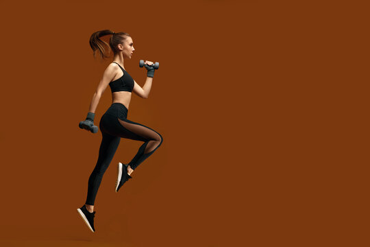 athletic beautiful woman in black sportswear training with dumbbells isolated on yellow background. girl jumping with dumbbells. sport and fitness concept. copy space