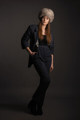 beautiful girl in a black trouser suit and a fur hat on her head posing in the studio on a dark gray background
