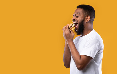 Black Millennial Guy Biting Burger Eating Over Yellow Background, Side-View