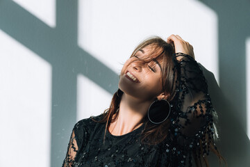 Happy female portrait. Boho stylish look. Party celebration. Smiling woman in black paillette decorated dress posing on neutral wall window sun light shadows .