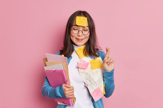 Positive schoolgirl crosses fingers believes in good luck on exam wears round spectacles stuck with papers and sticky notes written information to remember makes crib. Student uses cheat sheets.