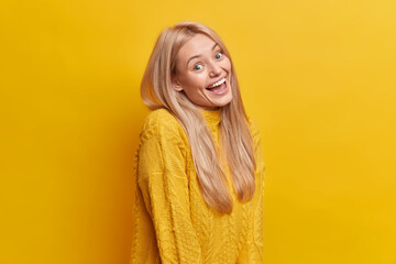 Plyaful cheerful blonde woman stands in profile looks gladfully at camera smiles toothily feels very happy wears casual jumper poses against vivid yellow background. People and emotions concept