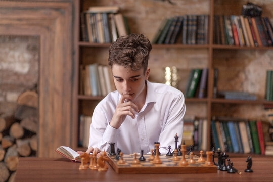The teenager plays chess with himself, the boy sits at a large wooden table and ponders chess moves in the game, white and black chess pieces, chess and checkmate.
