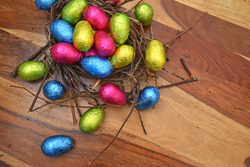 Foil wrapped colourful easter eggs in pink, green, blue and yelow in a natural nest made of sticks and twigs, against a multi grain brown wooden background.