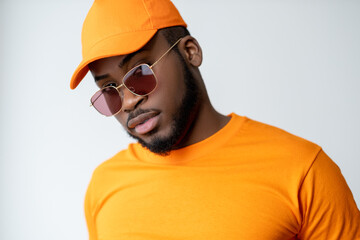Man fashion. Modern eyewear. Summer look. Confident cool African male model in orange t-shirt matching cap red color lens sunglasses looking at camera isolated on light neutral background.