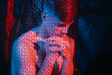Depressed man. Social inequality. Self isolation loneliness. Art portrait of hopeless thoughtful shirtless guy praying in mesh trap in red blue neon light at night on blur dark background.