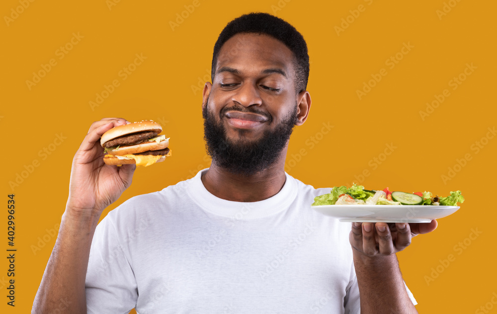 Wall mural Hungry Man Choosing Between Burger And Salad On Yellow Background