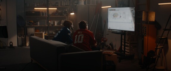 Two friends playing hockey sport video game inside garage hideout, enjoying pizza and drinks. TV...