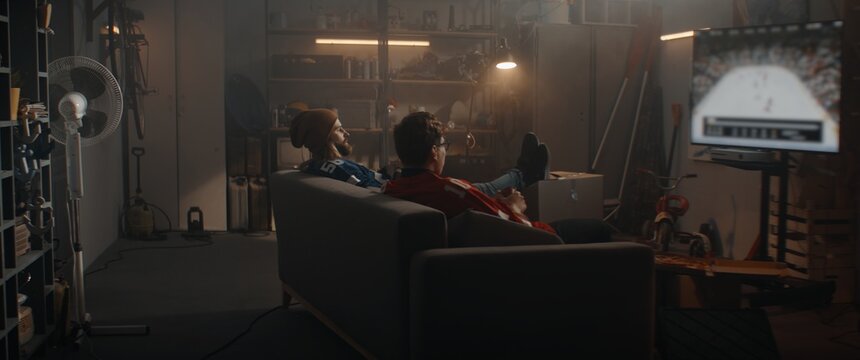 Two friends playing hockey sport video game inside garage hideout, enjoying pizza and drinks. TV screen is blurred
