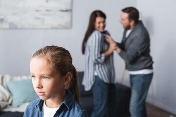 Upset kid standing near parents quarreling on blurred at home