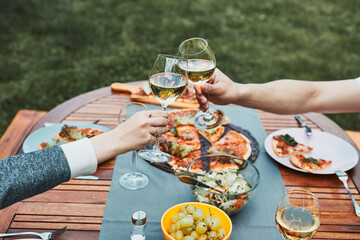 Friends making toast during summer picnic outdoor dinner in a home garden. Close up of people holding wine glasses with white wine over table with pizza, salads and fruits. Dinner in a orchard