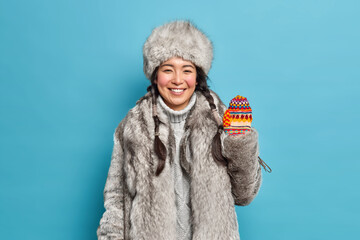 Positive brunette young Asian woman wears fur coat and hat waves hand in knitted mittens smiles...