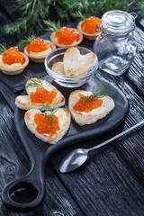 appetizer, heart-shaped snacks with butter and red caviar served with dill on a black wooden table