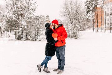 Young romantic couple is having fun outdoors in winter. Snowing. Love and leisure concept. Enjoying spending time together. Heart with sequins. Two lovers are hugging and kissing in Saint Valentine's 