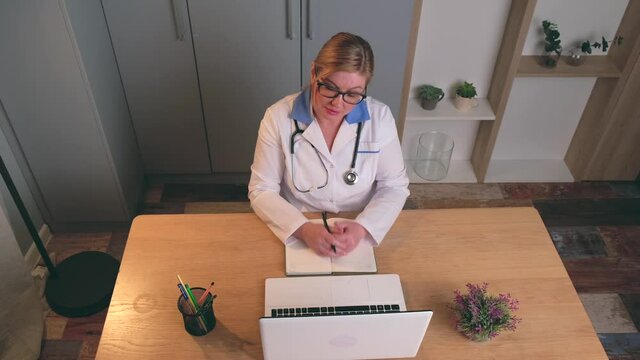 Caucasian Blonde Doctor With Glasses Is Sitting At The Laptop And Having A Video Meeting With A Patient Explaining The Next Steps Of The Cure Program