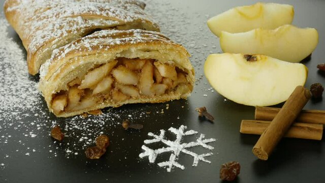 Puff pastry with apple filling. Apple strudel with cinnamon, raisins, cloves and powdered sugar snowflake. Rustic dessert with ingredients. Central european cuisine. 4k video, spinning table.